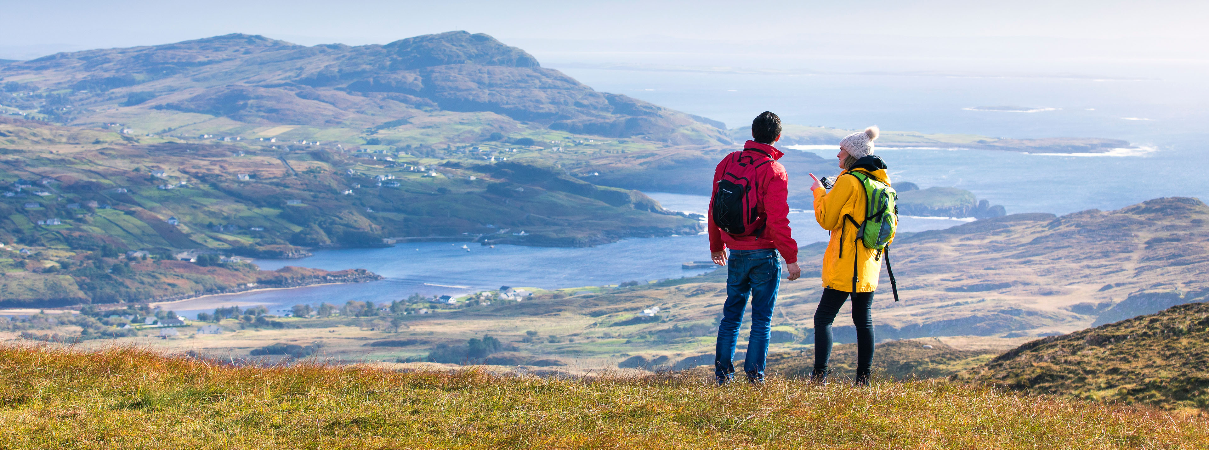 Two hikers overlooking a scenic Irish landscape with mountains and a shimmering lake in the distance.